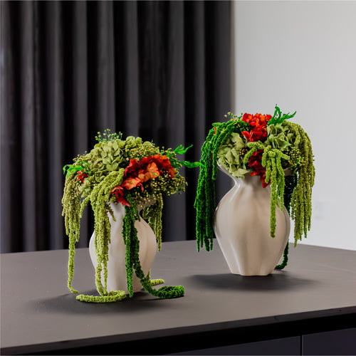 Zwei Blumensträuße mit ausgefallenen, hängenden Blumen stehen in sandfarbenen Keramikvasen mit fließenen Formen auf einer Anrichte