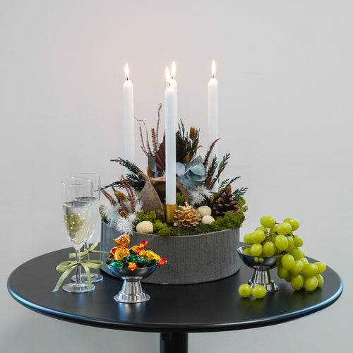 Auf einem Tisch stehen gefüllte Sektgläser, Süßigkeiten, grüne Trauben und eine Blumendekoration in einer großen Schale, in der vier Kerzen angezündet sind