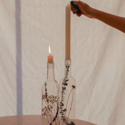 Auf einem Tisch stehen zwei Glasflaschen, die zu Kerzenhaltern umfunktioniert sind. In den Flaschen befinden sich getrocknete Blumen.