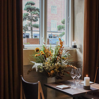 Vor einem Fenster steht ein hübsch gedeckter Holztisch mit einer brennenden Kerze und einem langlebigen Blumenstrauß mit warmen Farben, der eine gemütliche Atmosphäre schafft