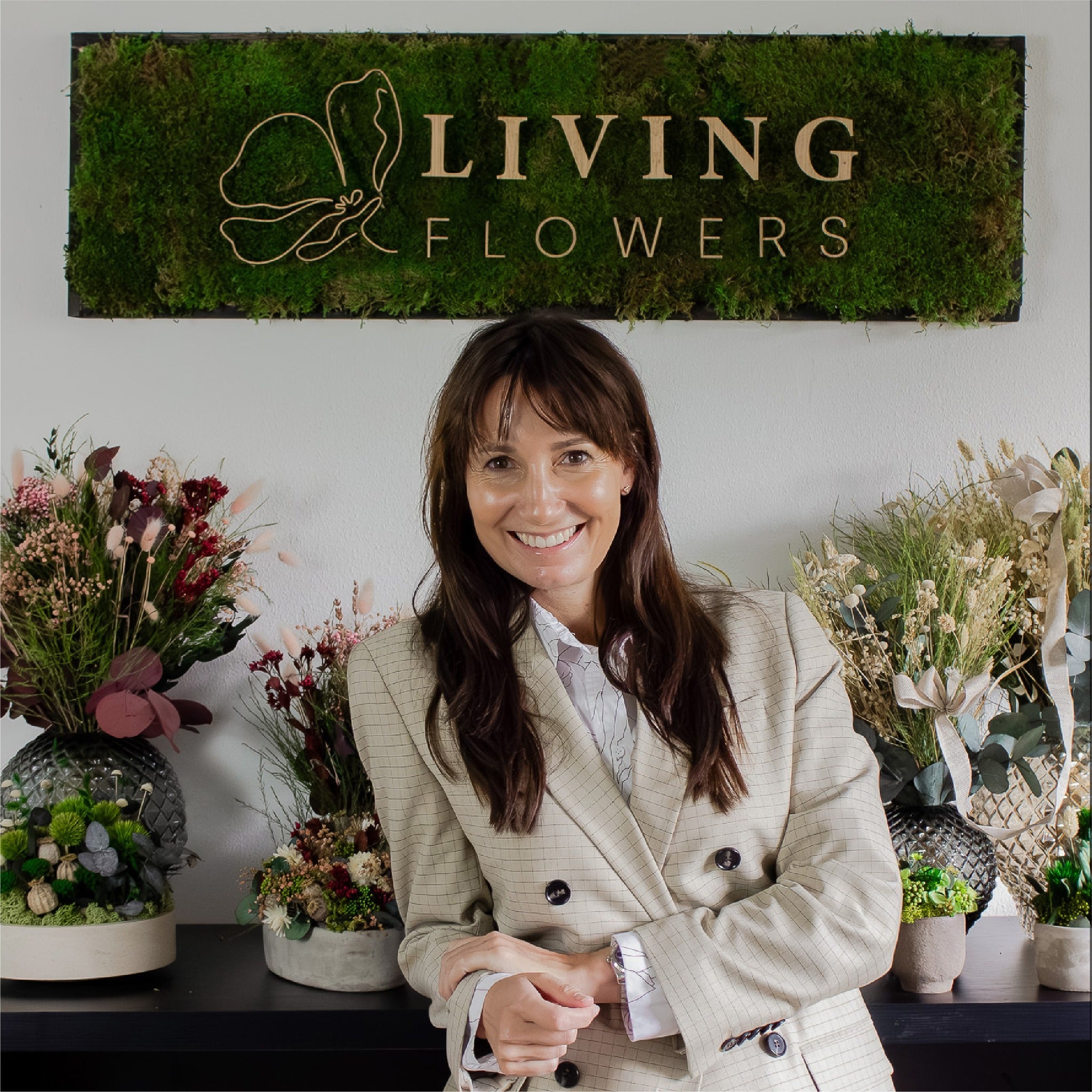 Die Gründerin von Living Flowers steht lächelnd vor einem Regal, auf dem langlebige Dekorationen und Blumensträuße stehen, über ihr hängt ein Schild mit der Aufschrift Living Flowers