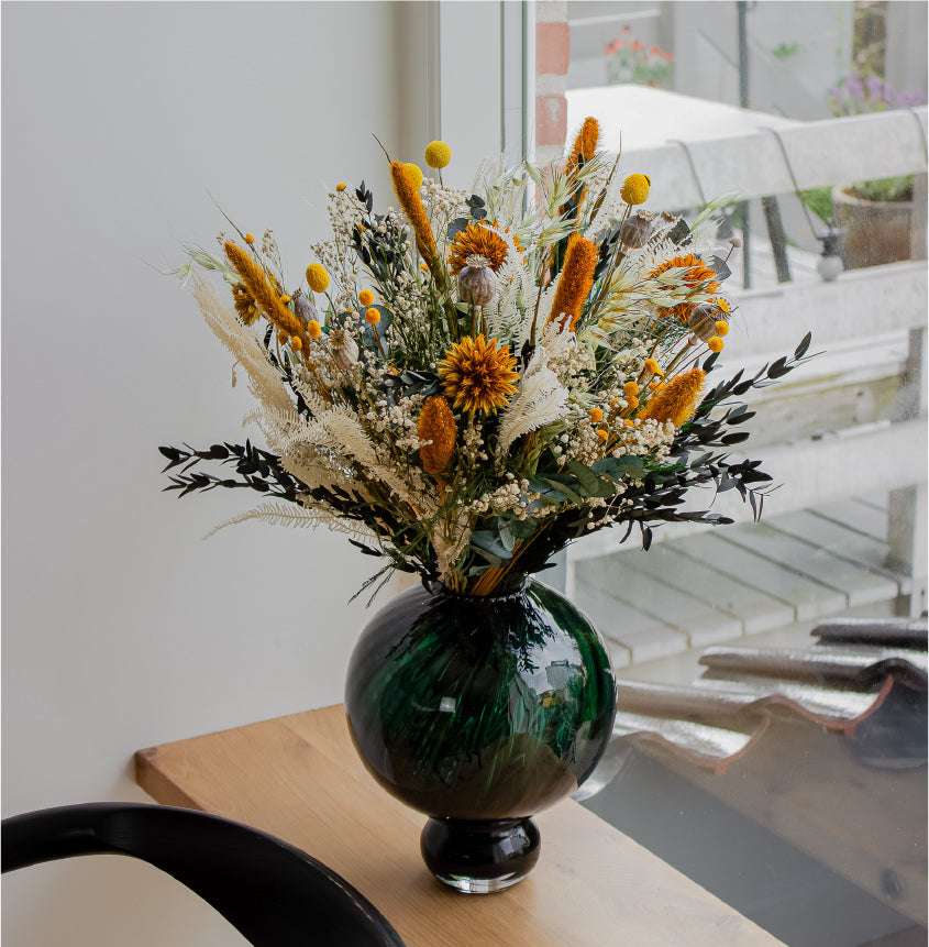Auf einer Fensterbank steht ein oranger Blumenstrauß in einer bauchigen grünen Vase