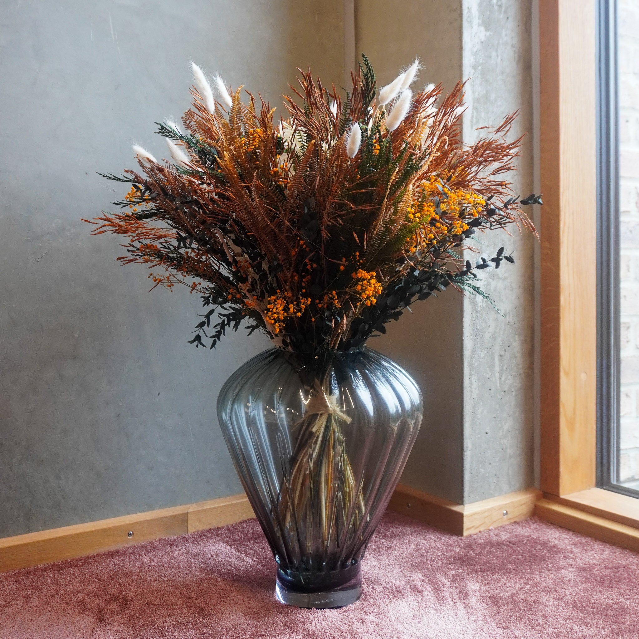 In einer Zimmerecke steht ein voller Blumenstrauß in warmen Herbstfarben in einer geometrischen Glasvase auf dem Boden