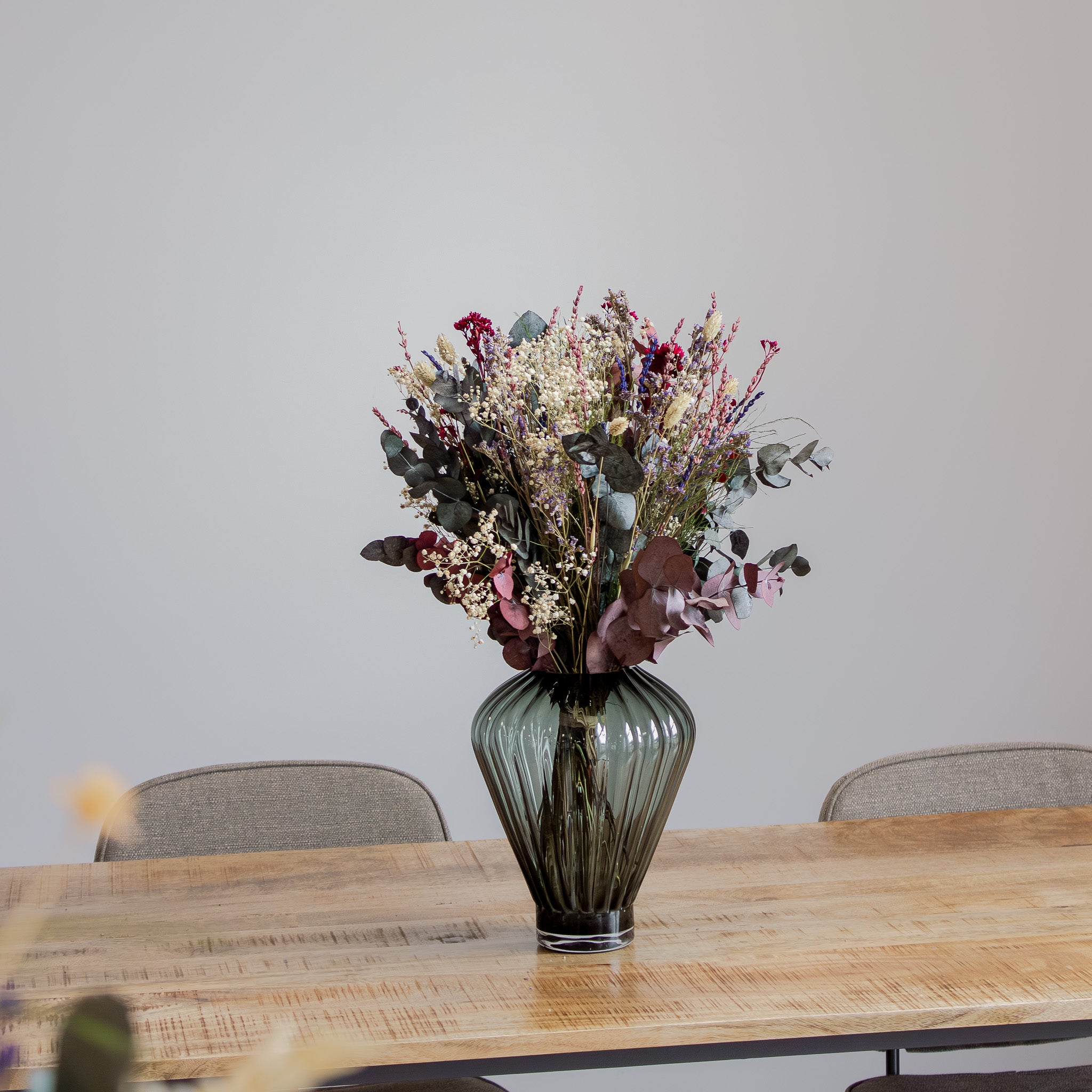 In einer geriffelten Glasvase auf einem Holztischt steht ein wilder, natürlicher Blumenstrauß mit violetten, blauen und roten Akzenten