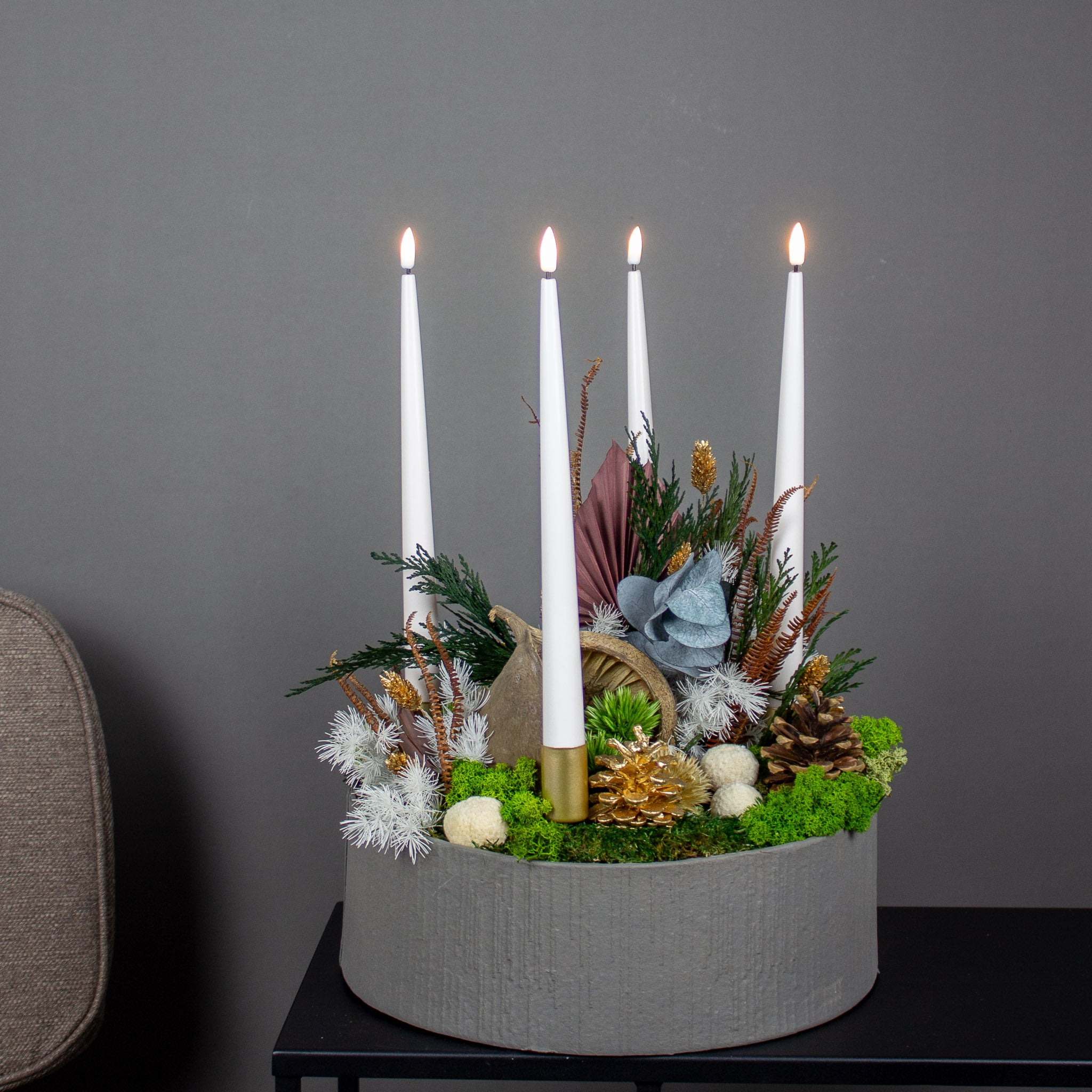 In einer Schale ist eine weihnachtliches Blumengesteck aus Moos, Tannenzapfen und langlebigen Blumen sowie vier Kerzen arrangiert