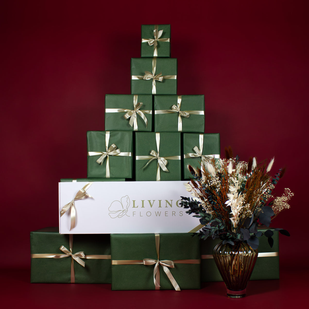 Vor einer Pyramide aus grünen Geschenken, in der auch eine weiße Living Flowers Box liegt, steht eine Glasvase mit einem weihnachtlichen Blumenstrauß