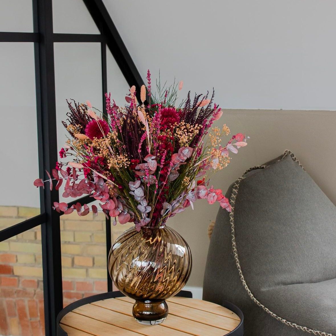 Ein Blumenstrauß in kräftigen Rot- und Pinktönen stehet in einer Vase auf einem Beistelltisch