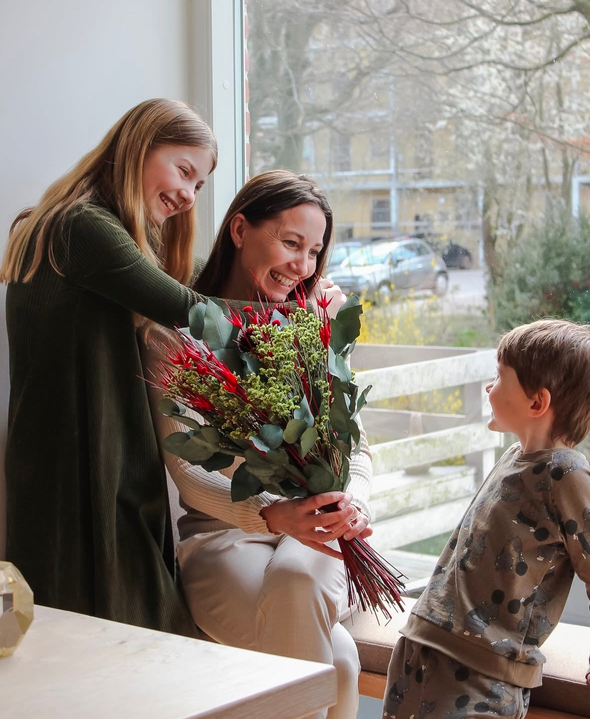 Eine Mutter sitzt mit ihren beiden Kindern in einem Wohnraum, alle lächeln. Die Mutter hält einen grün-roten Blumenstrauß