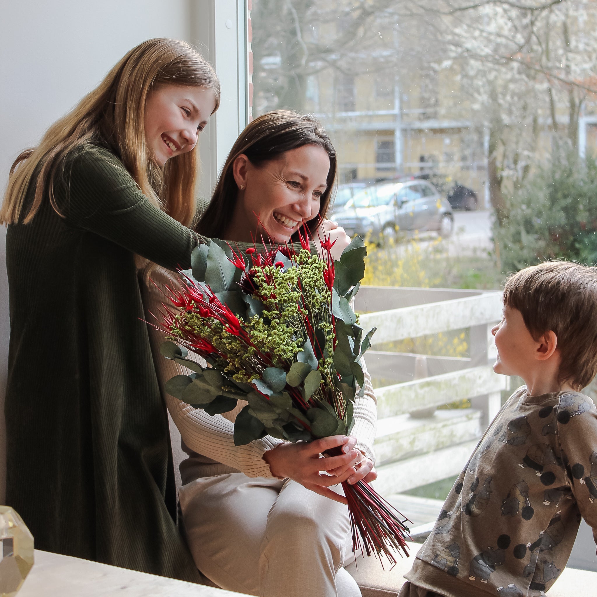 Eine Mutter sitzt mit ihren beiden Kindern vor einem Fesnter, sie hält einen grün-roten Blumenstrauß in der Hand, alle lächeln