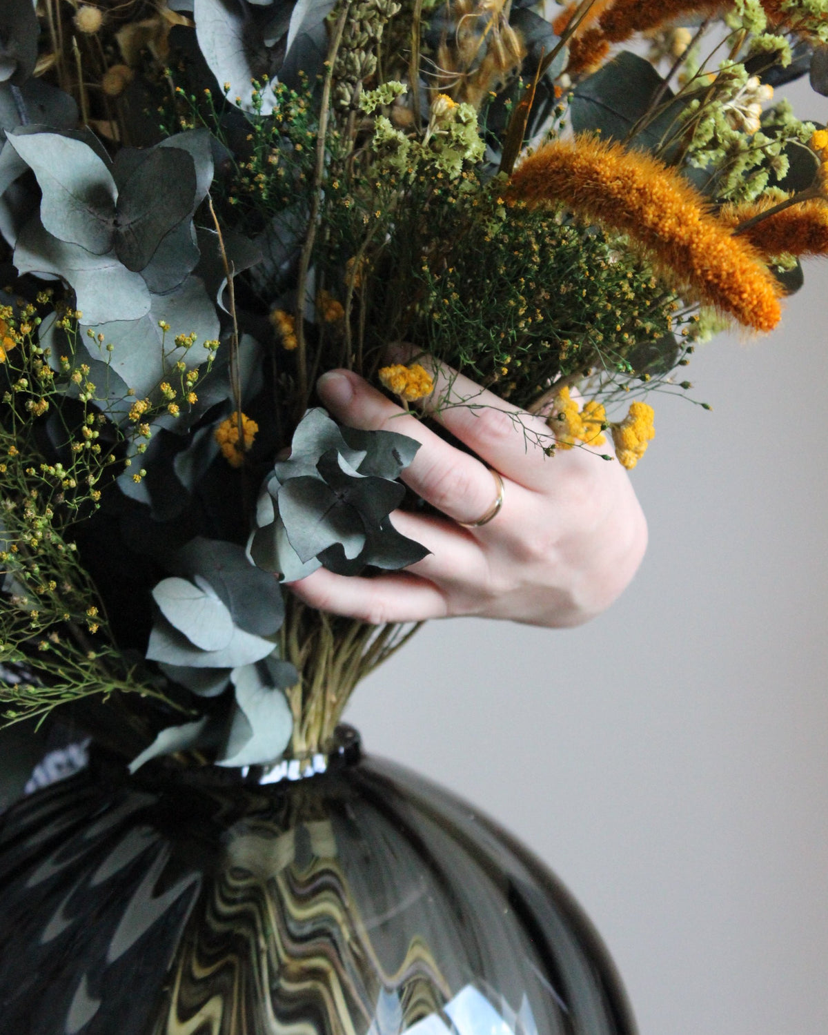 Nahaufnahme eines Blumenstraußesin warmen Herbstfarben, der von einer Frau in einer bauchigen Glasvase drapiert wird
