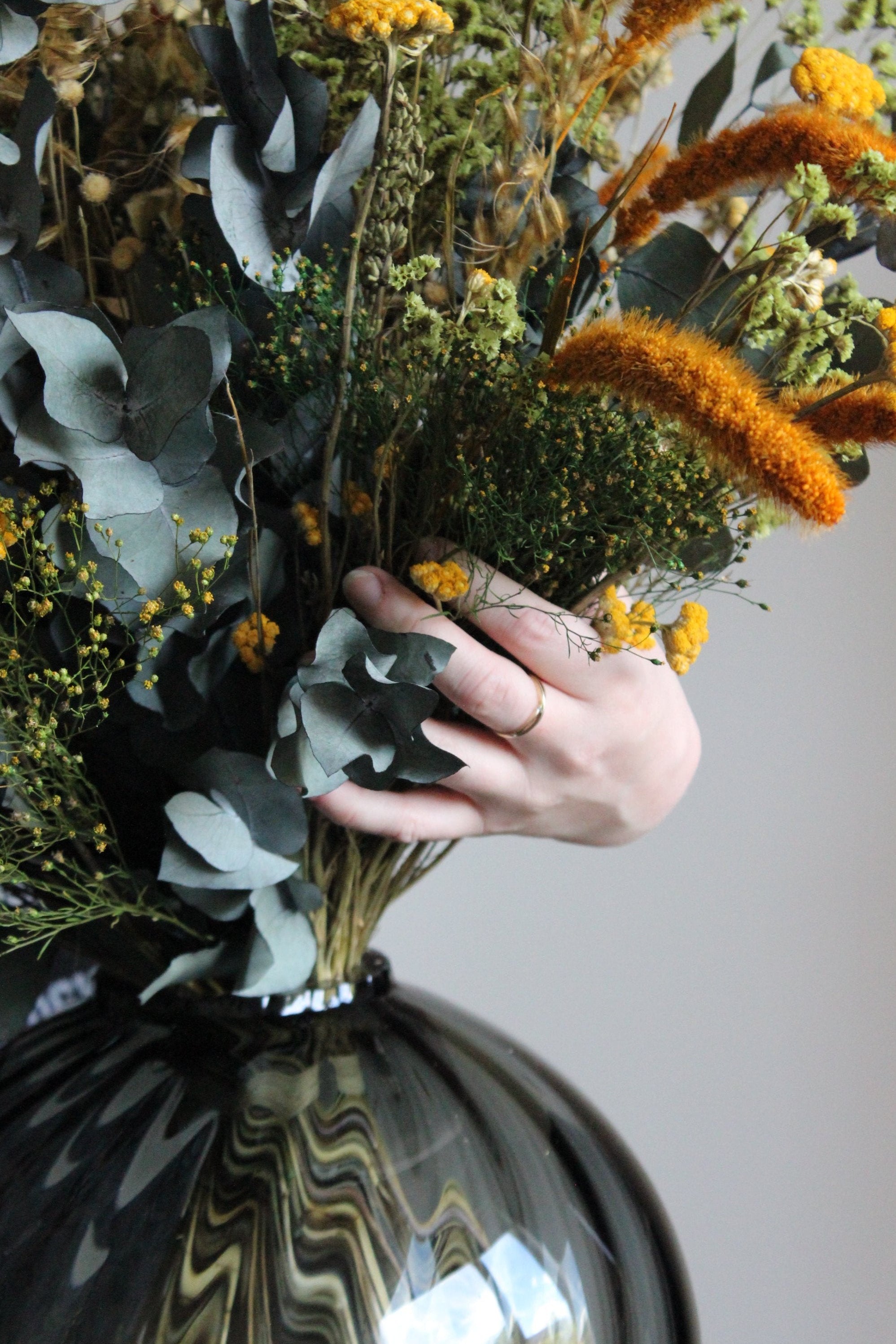 Nahaufnahme eines Blumenstraußesin warmen Herbstfarben, der von einer Frau in einer bauchigen Glasvase drapiert wird