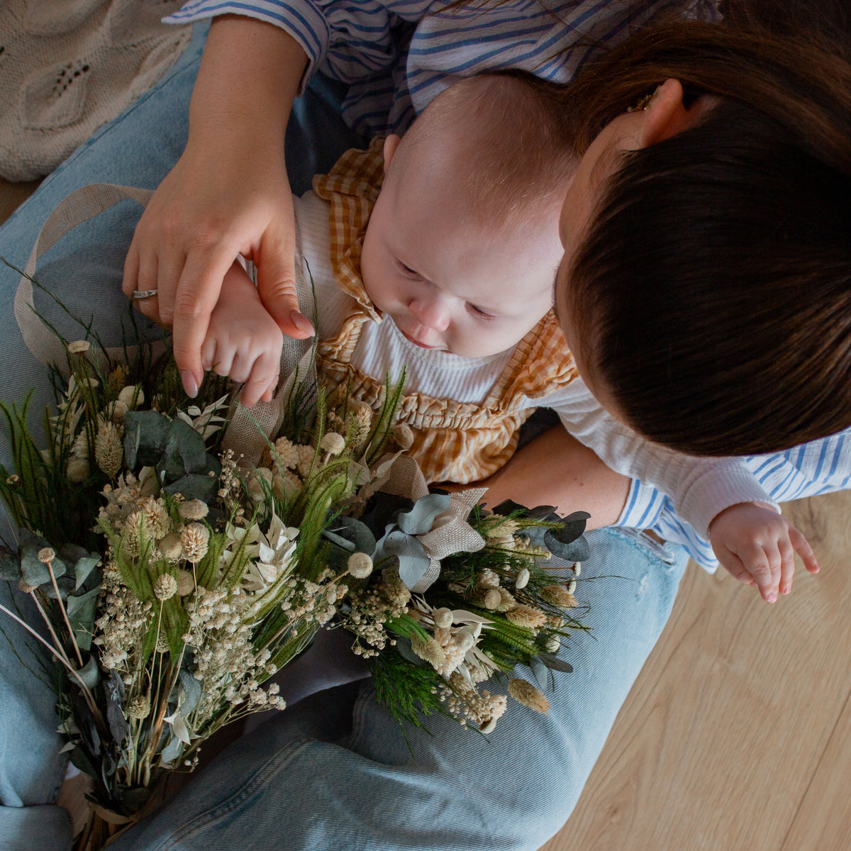 Et barn sidder på sin mors skød og rører ved to små, lyse blomsterbuketter.