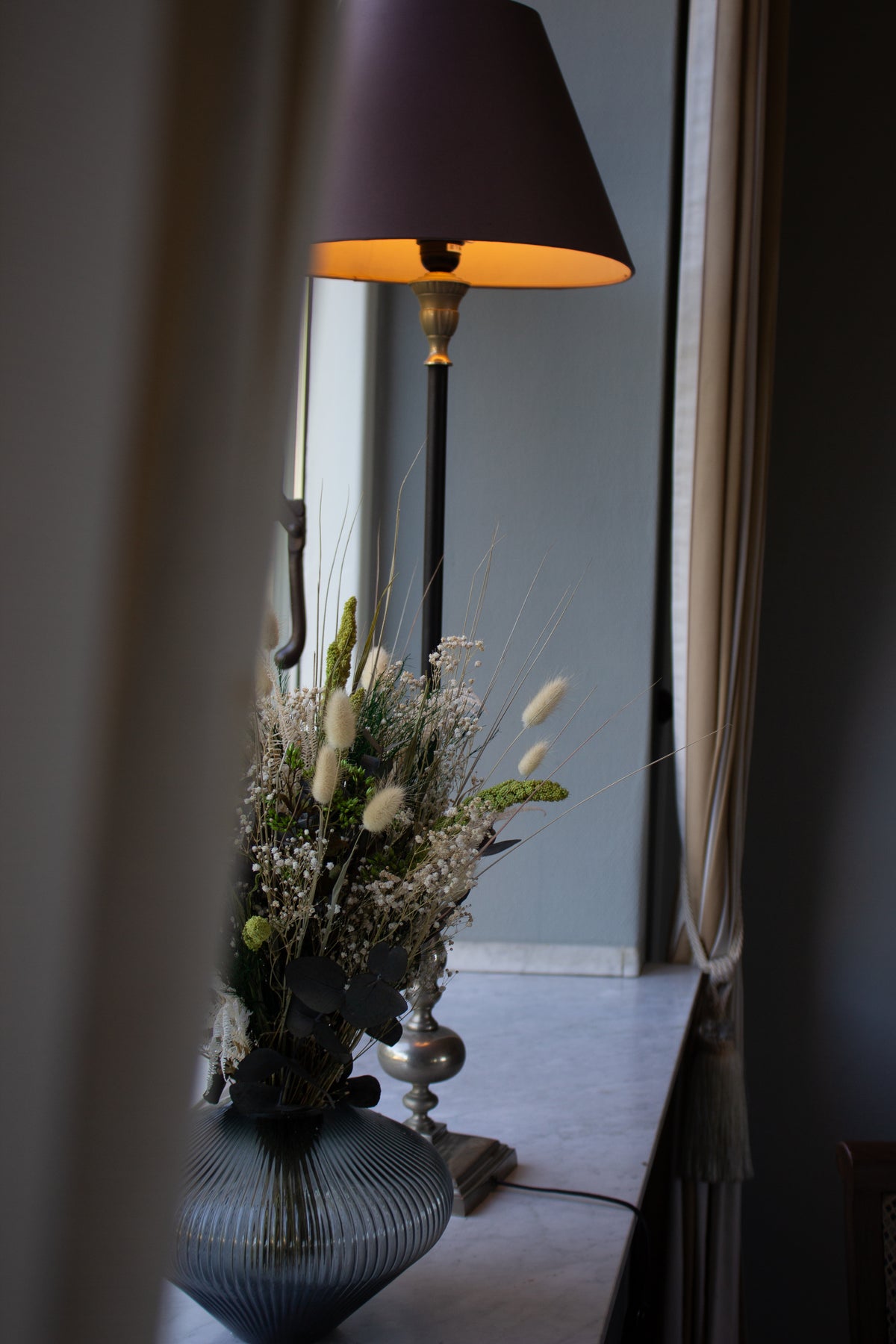 Auf einer Fensterbank steht neben einer altmodischen Lampe ein grün-weißer Blumenstrauß in einer Glasvase
