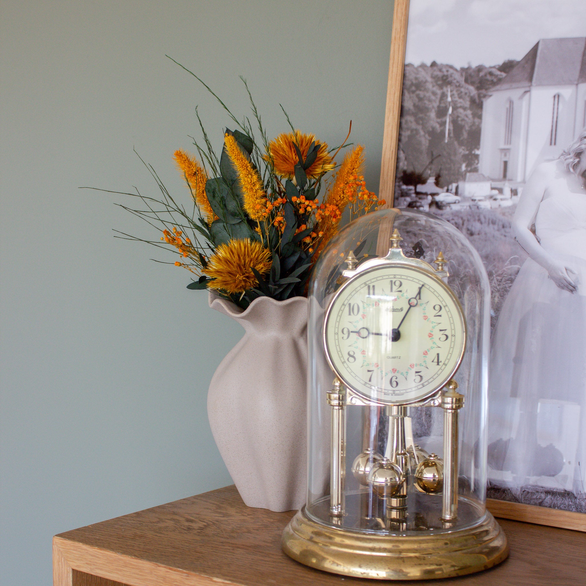 Auf einem Regal steht neben einer antiken Uhr ein Miniaturblumenstrauß in kräftigem Orange in einer sandfarbenen Vase