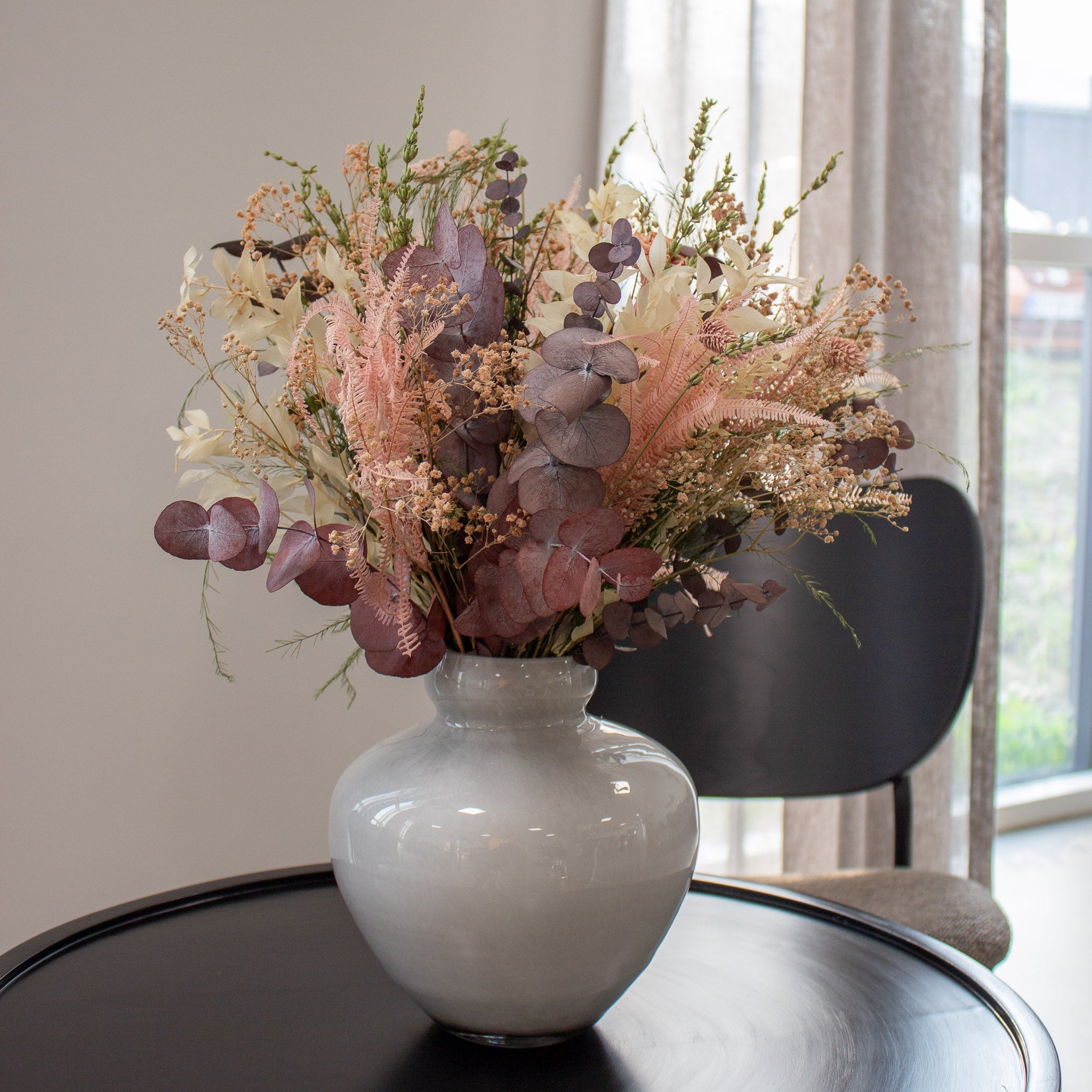 Auf einem Tisch steht eine Vase bauchige Vase aus weißem Milchglas, in der ein zarter rosafarbener Blumenstrauß steht.