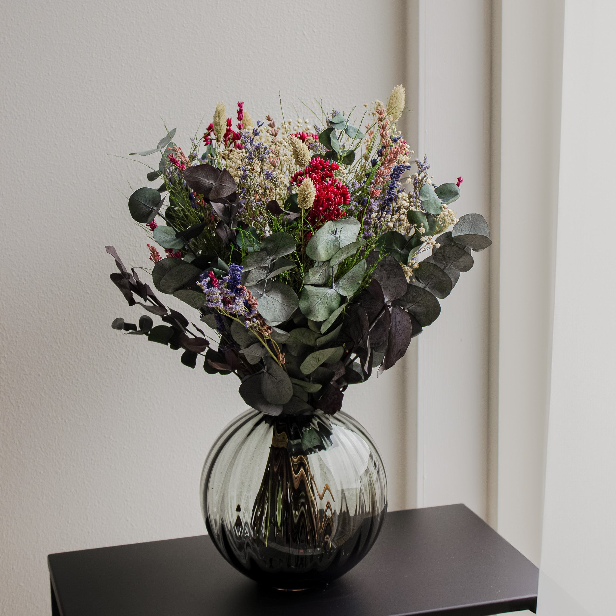 Auf einem kleinen Beistelltisch steht ein natürlicher Blumenstrauß mit violetten, blauen und roten Akzenten in einer bauchigen Vase