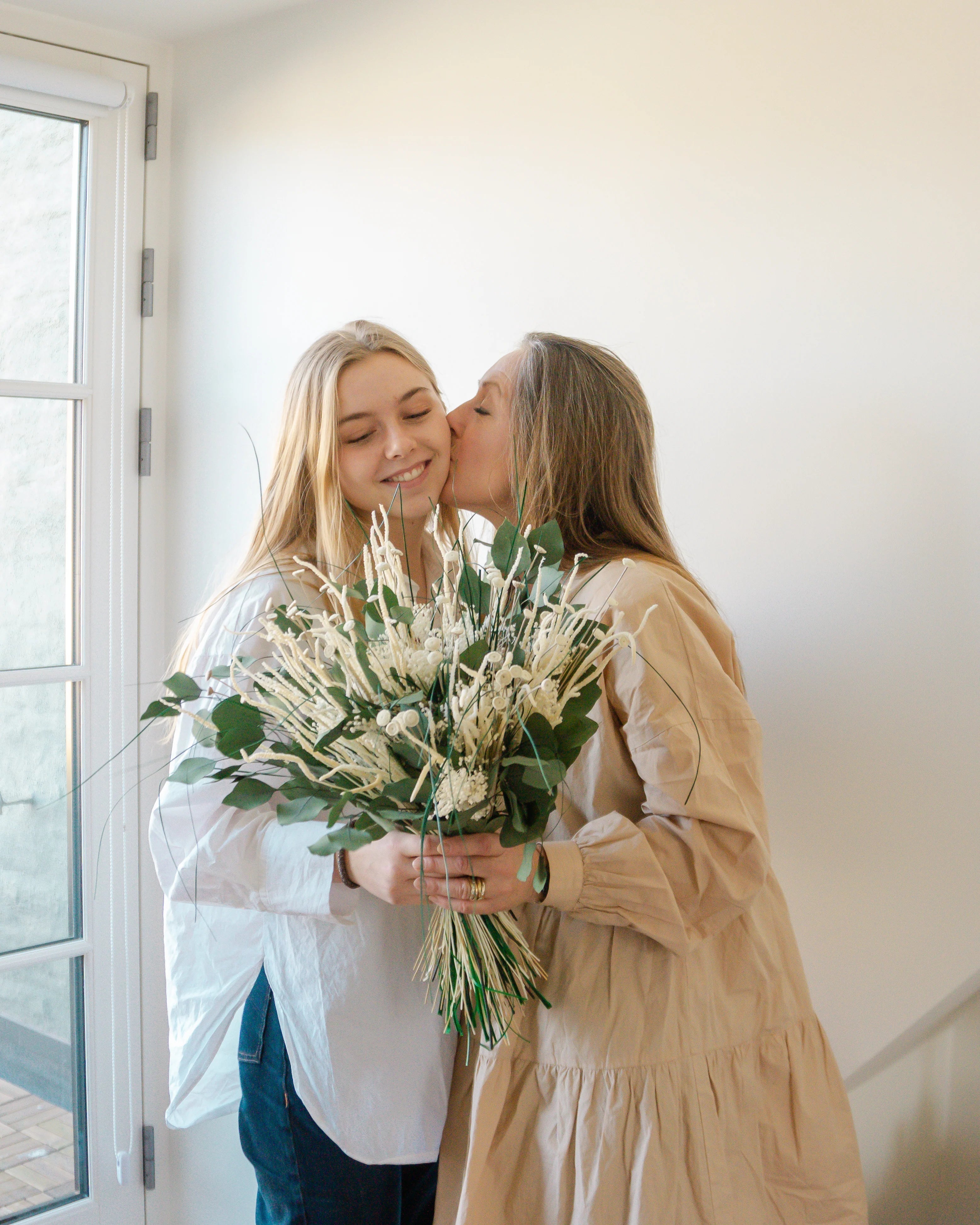 Eine Tochter reicht ihrer Mutter einen weiß-grünen Blumenstrauß, die Mutter küsst sie auf die Wange