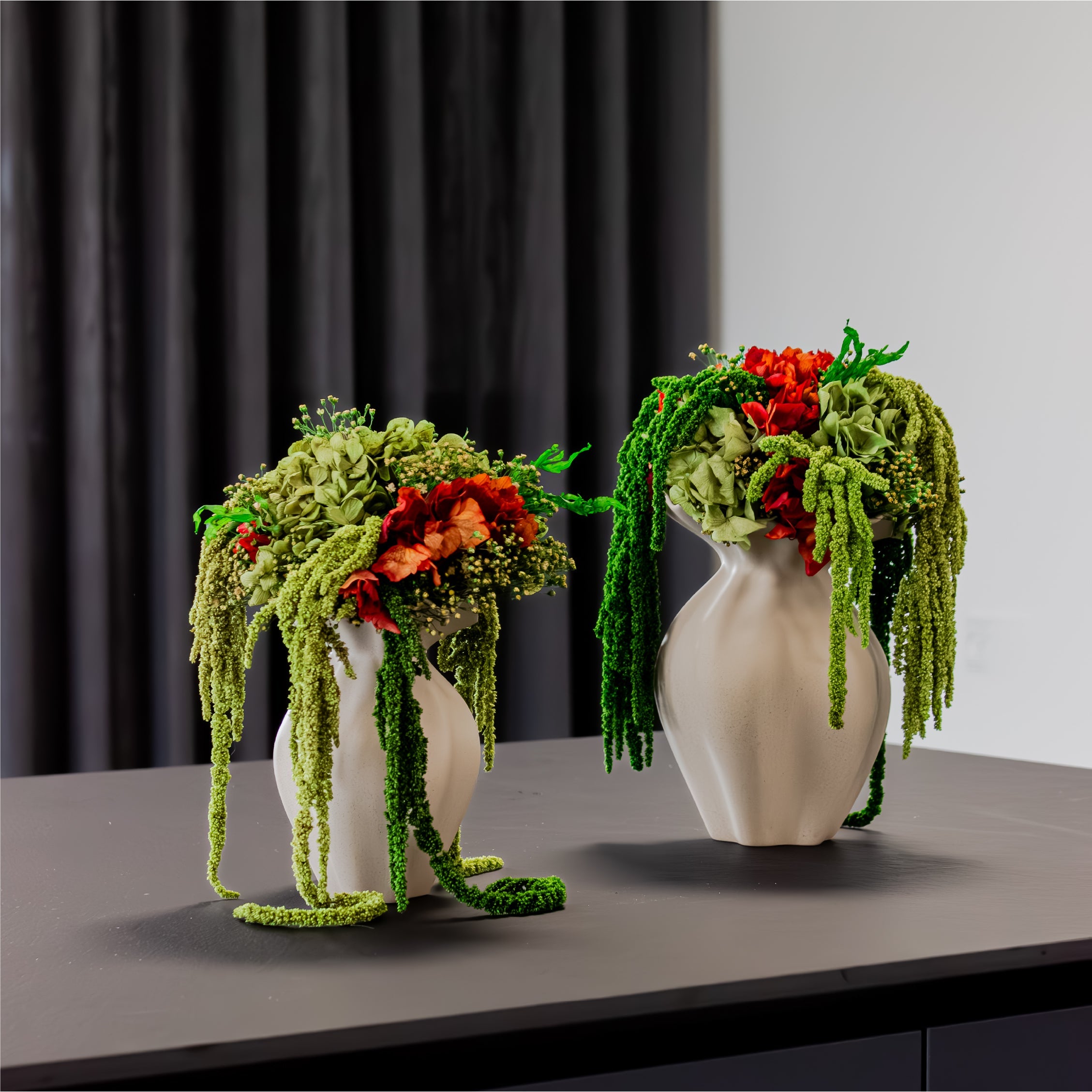 Auf einem Tisch stehen zwei aufwändig gebundene grün-orange Blumensträuße mit hängenden Elementen in sandfarbenen Keramikvasen 
