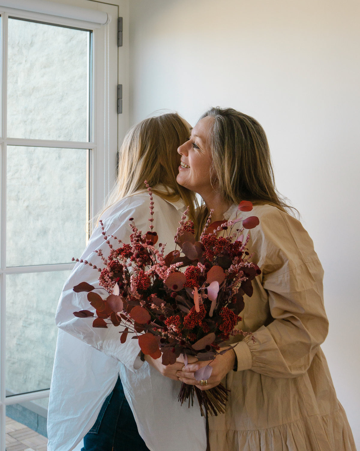 En datter giver sin mor en stor blomsterbuket i røde nuancer, og de krammer hinanden.