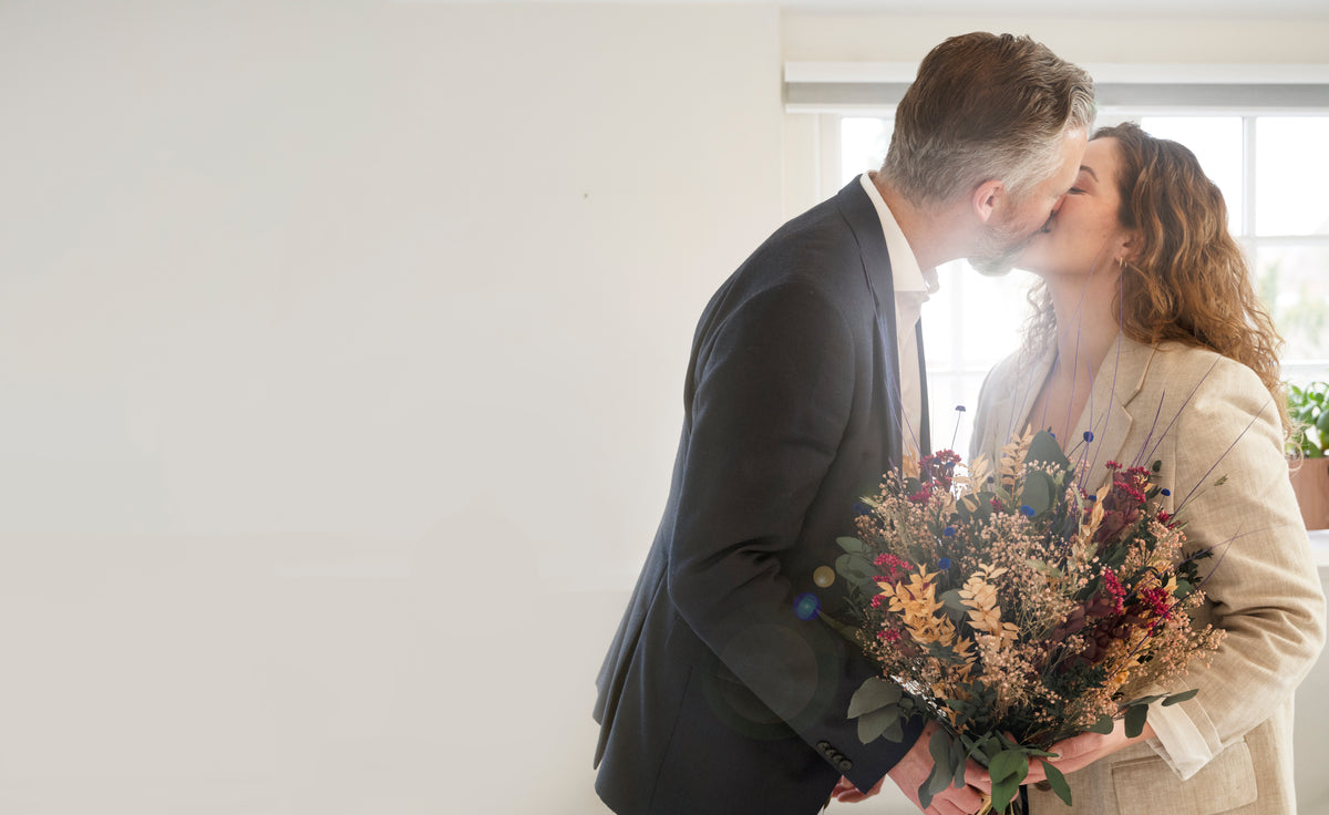 Paar küsst sich und hält einen langlebigen Living Flowers Blumenstrauß mit roten, orangen und blauen Blüten sowie Eukalyptus.