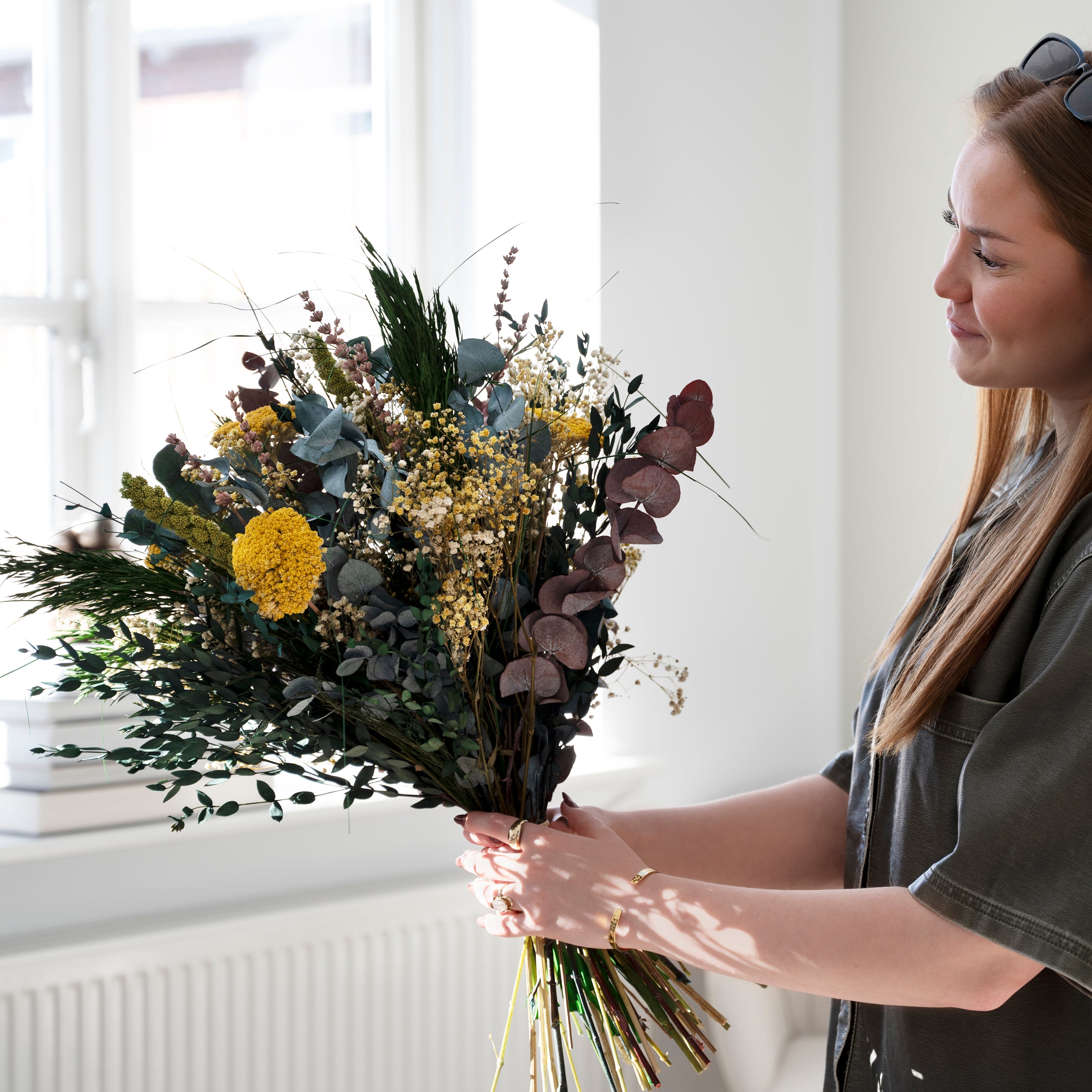 Frau hält den Living Flowers Strauß Sonnenstrahl mit Eukalyptus, gelben Blüten und dunkelroten Blättern vor einem hellen Fenster
