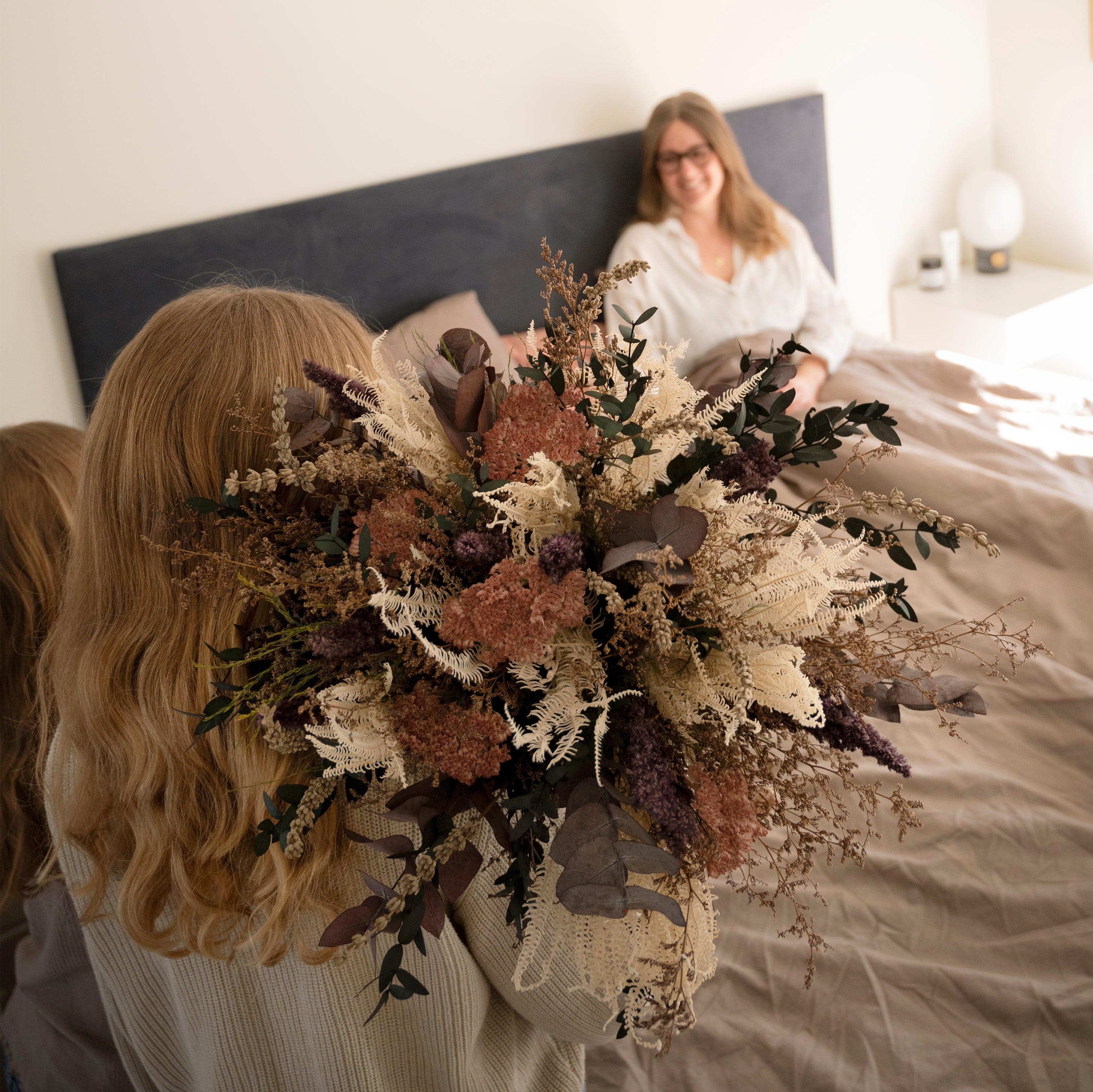 Kind überreicht Mutter einen großen Living Flowers Blumenstrauß mit rosafarbenen, lila und cremefarbenen Blüten sowie Eukalyptus im Schlafzimmer.