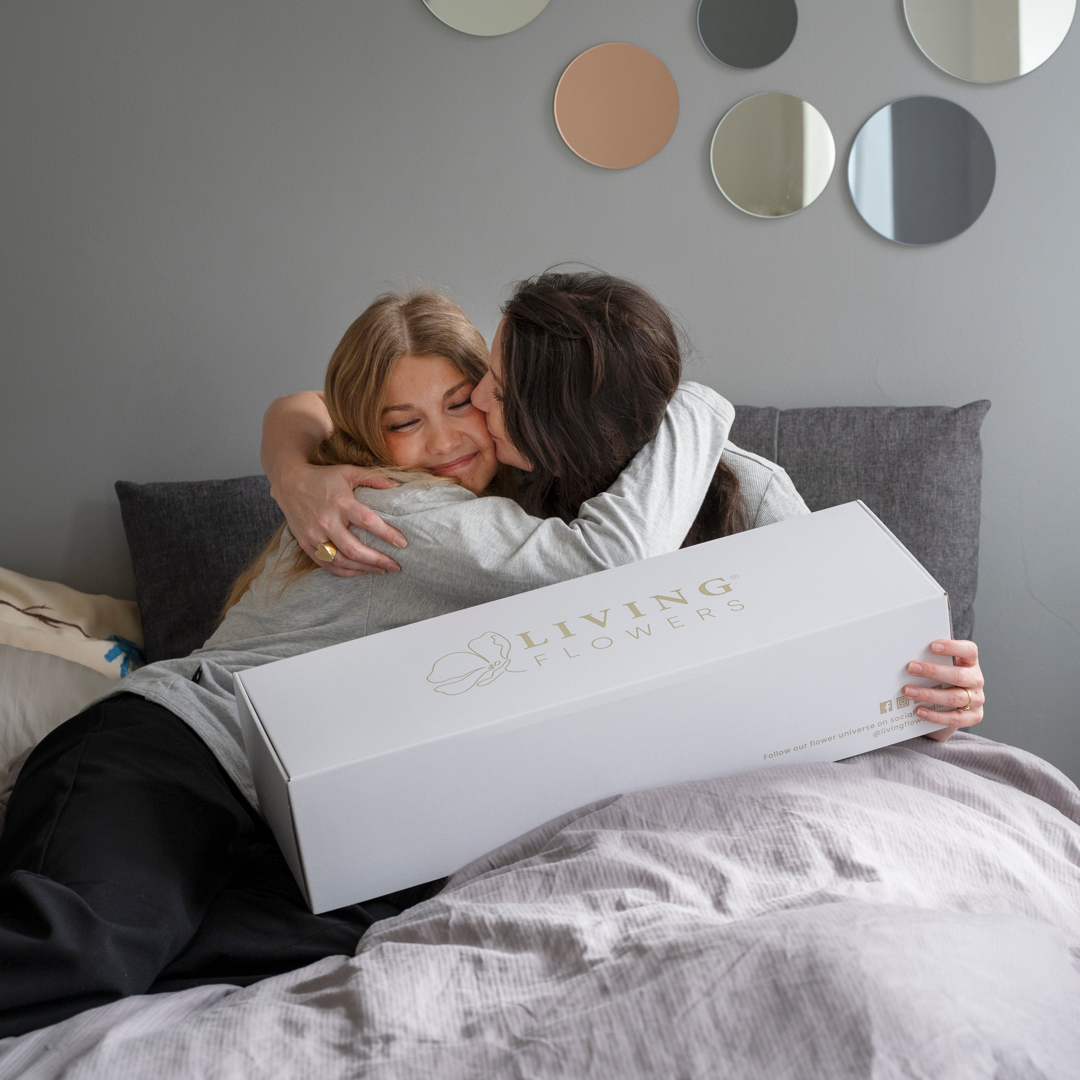 Zwei Frauen umarmen sich freudig auf dem Bett mit einer geöffneten Living Flowers Geschenkbox in der Hand