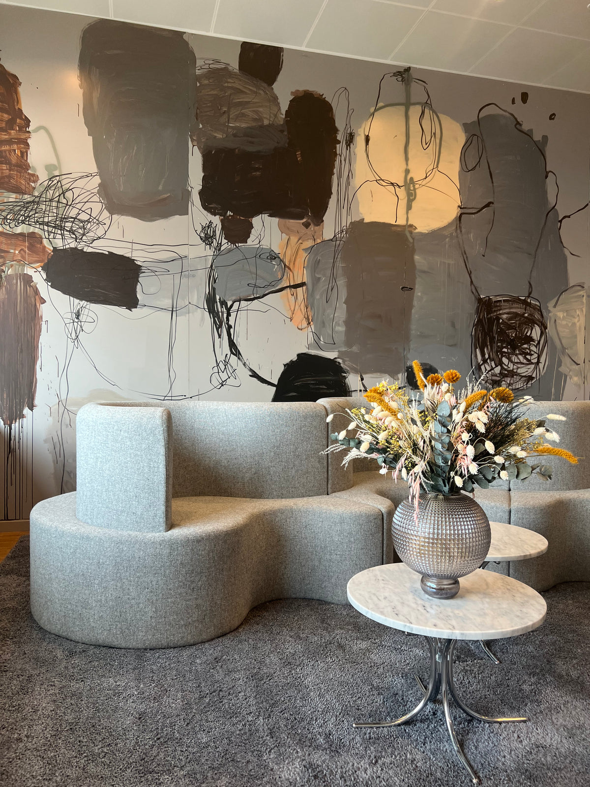 På et lille sidebord i et excentrisk indrettet rum står en stor, buet blomster-vase med en orange-hvid blomsterbuket.