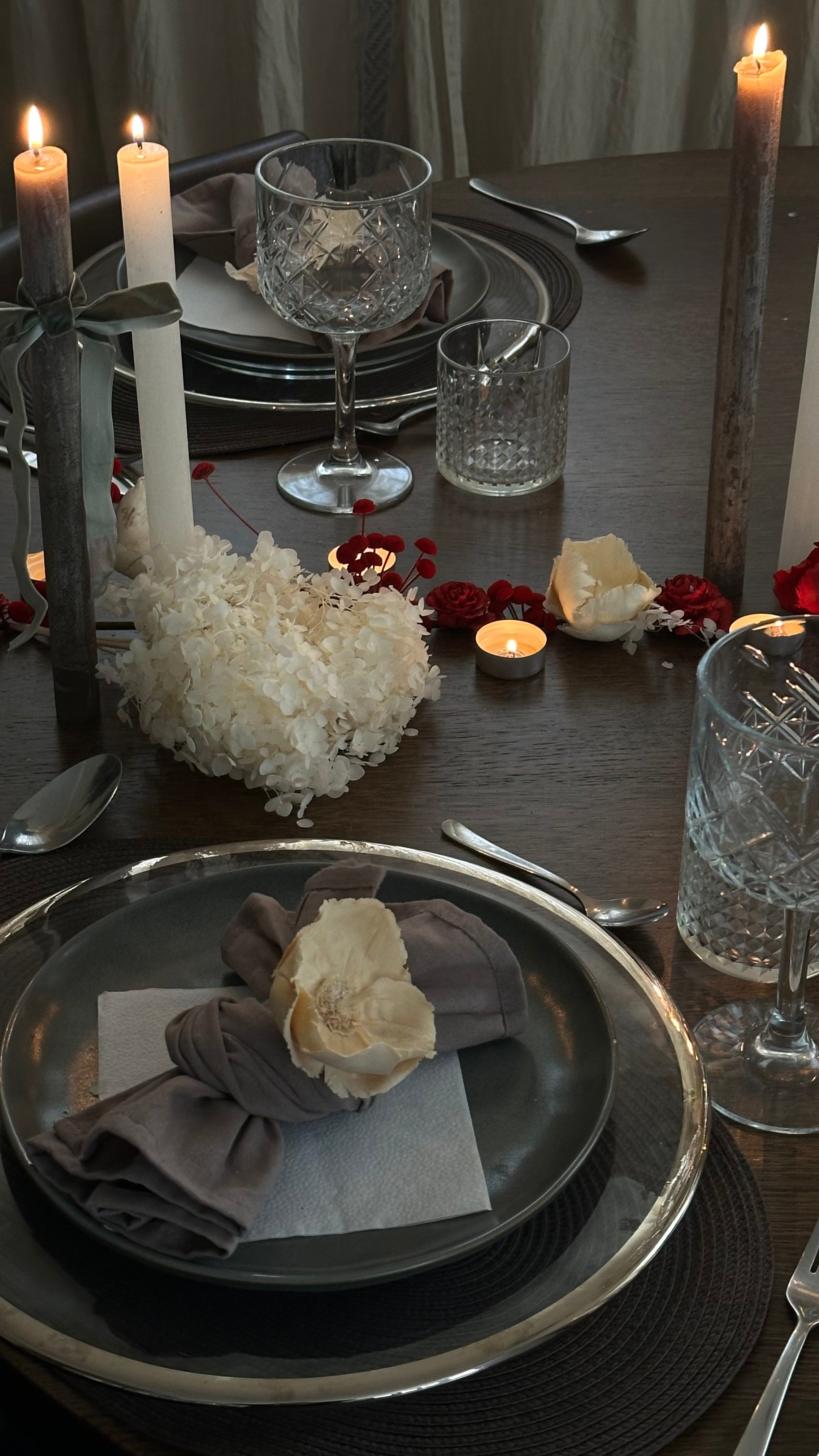 Et stemningsfuldt og elegant dækket bord med mørke toner, levende lys, krystalglas og en blomsterdekoration i rødt og hvidt – perfekt til en romantisk aften.