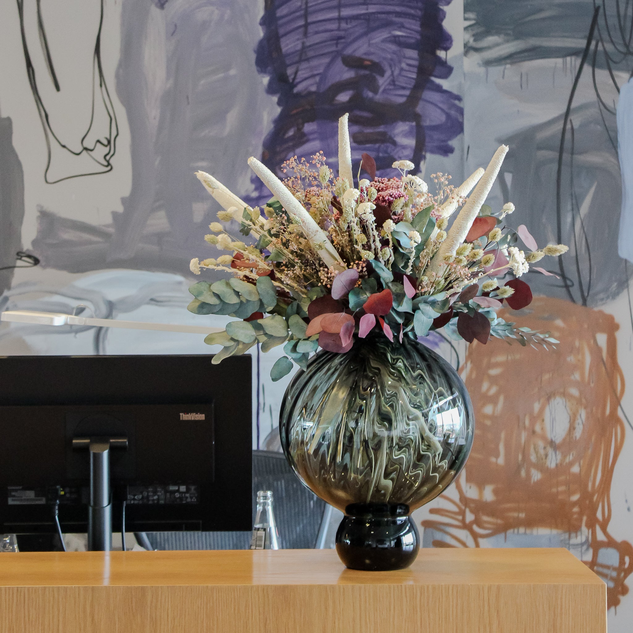 Auf dem Tresen des Hotels steht eine bauchige Vase mit einem ausdrucksstarken Blumenstrauß mit unterschiedlichen Eukalyptussorten und weißen Akzenten