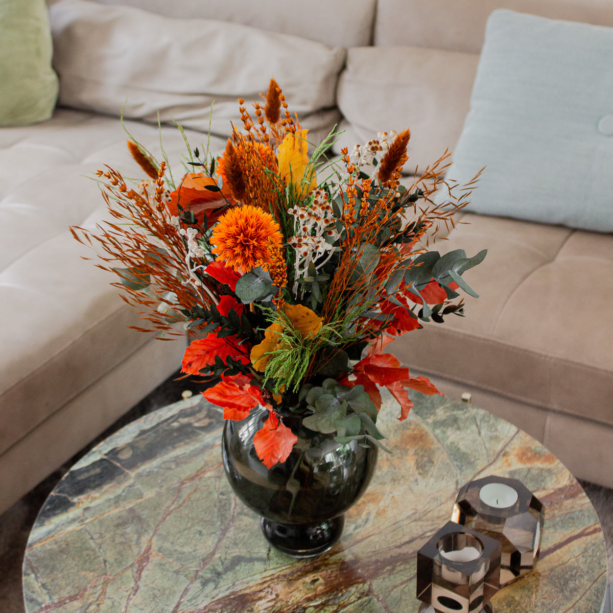 Bukket med langtidsholdbare blomster i varme efterårsfarver står i en mørkegrøn, buet vase på et marmorbord foran en beige sofa.