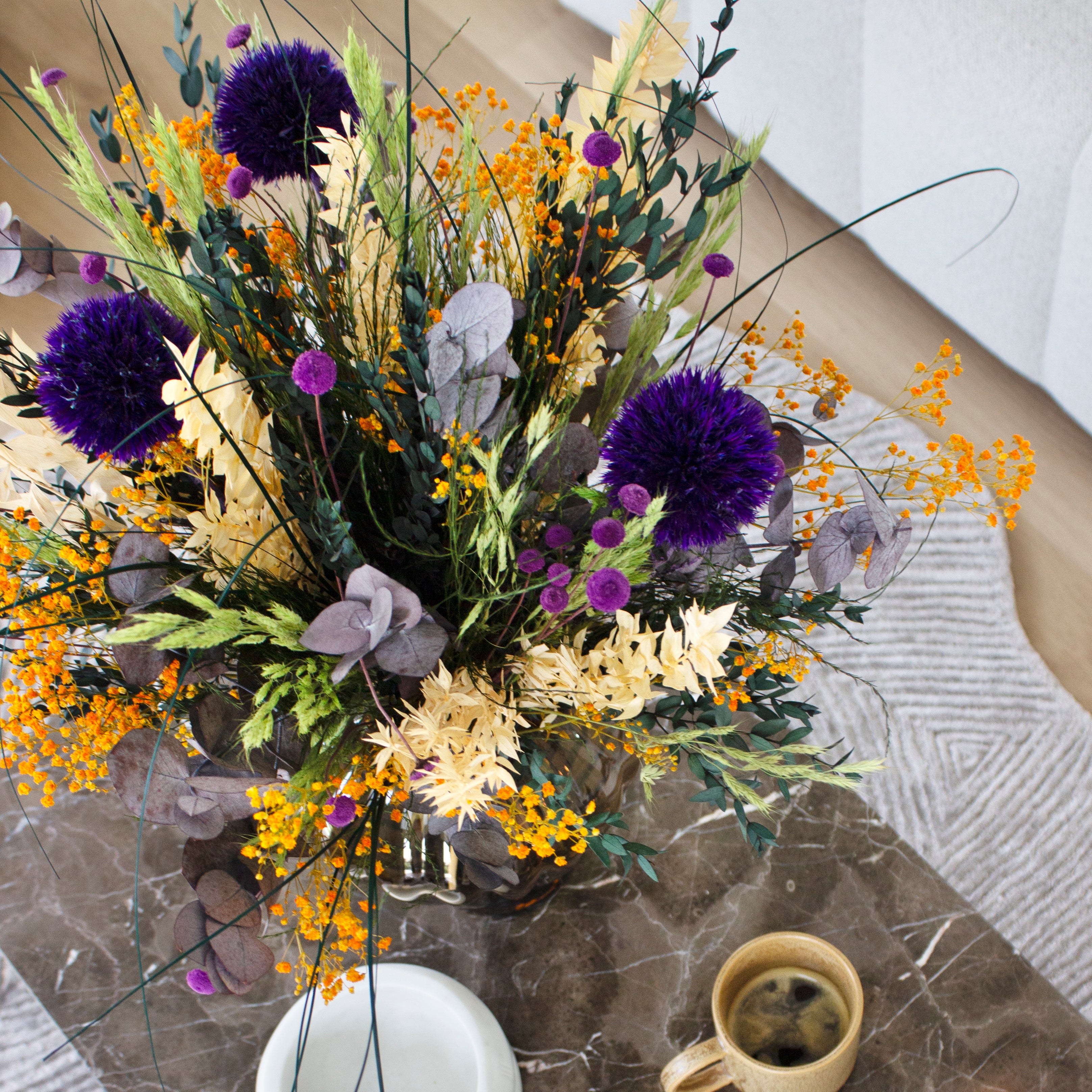 Bunter Blumenstrauß mit violetten Kugelblüten, gelben und orangefarbenen Akzenten sowie Eukalyptus auf einem Marmortisch neben einer Kaffeetasse