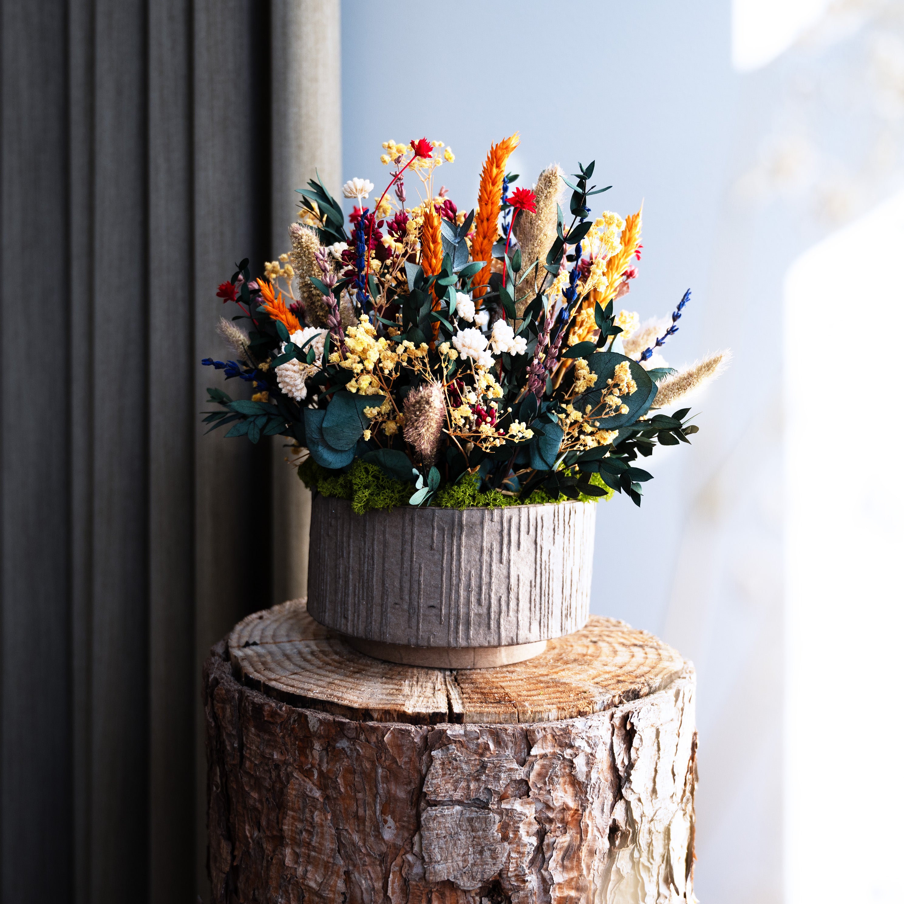 Langlebiges Blumenarrangement mit orangefarbenen, roten und cremefarbenen Blüten sowie Eukalyptus in einem dekorativen Topf auf einem Holzstamm