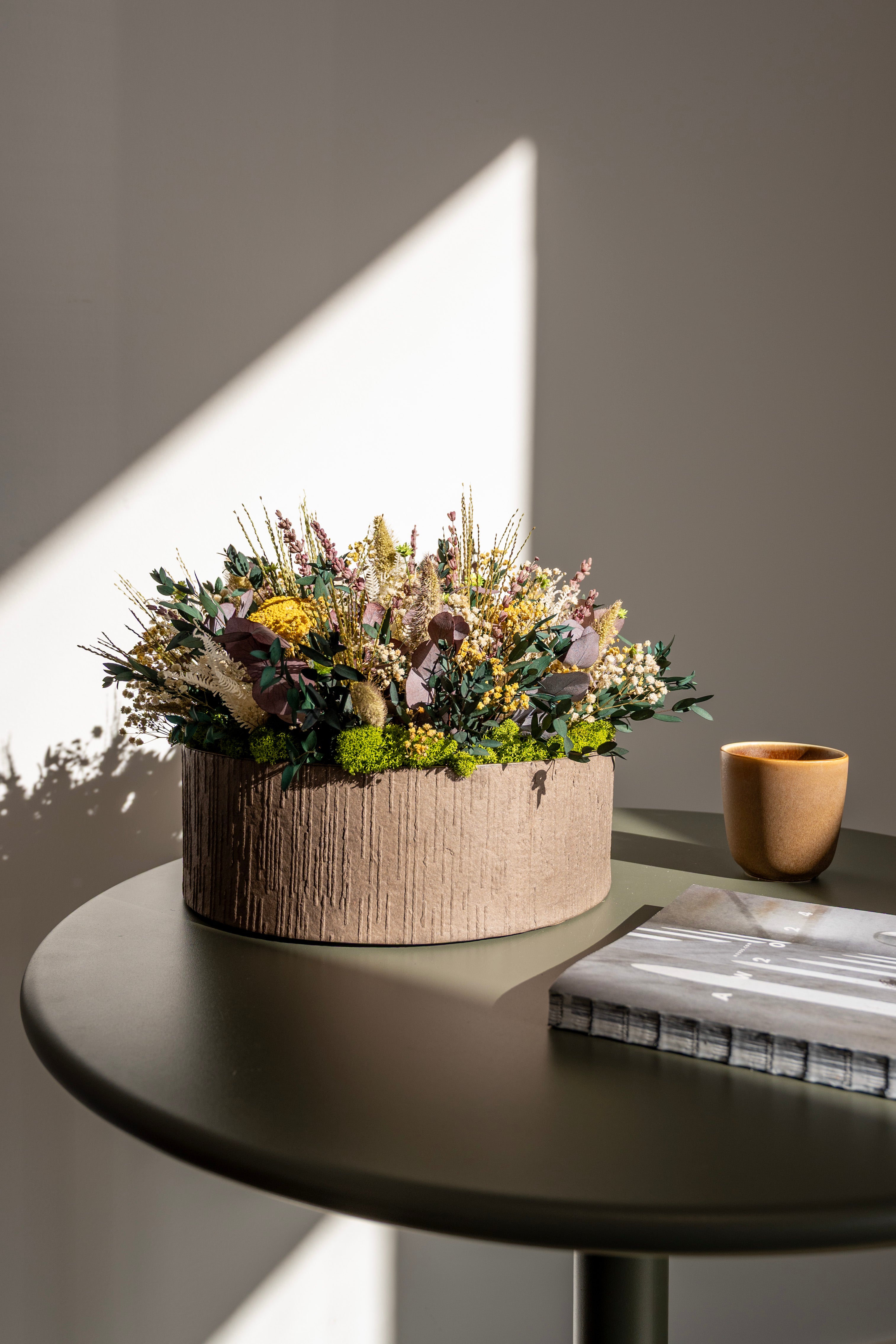 Langlebige Living Flowers Blumendekoration mit gelben, lila und grünen Blüten in einem runden Rindentopf auf einem dunklen Beistelltisch